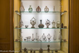 Musée des Arts Décoratifs. Chambre Jonquille.<br> Etagère supérieure carafes, gourdes, décor à la pince, décor de filets rudentés et décor de pastilles de verre France. XVIIIe siècle.
La lampe à huile utilisée par les dentellières s'accompagnait d’un globe rempli d'eau faisant loupe.
Deuxième étagère :
De gauche à droite : Bénitier en verre blanc, décor à la pince, France, XVIIIe siècle,
Tasse à via à anse à décor de filets bleus à la pince, Nevers, XVIIe ou XVIIIe siècle.
Deux cantirs de baptême, Catalogne, XVIIIe siècle.
Au centre, gourde en verre tacheté bleu-vert, rouge et blanc, Montagne-Noire ou Nevers, 1e moitié du XVIIe siècle ; vase d’aspersion d'eau de rose dite alnorraxa, Catalogne. XVIIe siècle, cruche à décor de filets blancs peignés, Espagne, fin XVIIIe siècle, et flacon mauve à décor peigné. Catalogne ou Languedoc, XVIIIe siècle.
Troisième étagère :
De gauche a droite, veilleuse, France, XVIIIe siècle, huilier-vinaigrier, France, XVIIIe siècle, fuseau a dévider en verre mauve, Sud-Ouest ou France, XVIIIe siècle ; deux flacons d'amusement en forme d'animaux, hérisson et souris, XVIIIe siècle, deux essenciers et un essencier ou biberon en verre blanc. Sud de la France, fin du XVIIIe siècle; deux cruches de table, Normandie et Sud-Est, XVIIIe siècle, petite burette façon de Venise, France, XVIIe siècle, verre à coupe conique façon de Venise, Pays-Bas, XVIIe siècle : bouteille essencier, France XVIIIe ou début XIXe siècle ; baromètre à eau piriforme, avec anneau de suspension, France XVIIIe.
Quatrième étagère :
A gauche, verres opalins de Bohème, milieu du XVIIIe siècle.
Une théière et un confiturier à décor peint de fleurs polychromes inspiré des motifs de la Compagnie des Indes. Deux moutardiers et deux gobelets ont un décor peint d'inspiration occidentale de style rocaille.
A droite, quelques exemples d'une production verrière française de la fin du XVIIe siècle par Bernard Perrot à Orléans et la Verrerie Royale, en activité de 1662 à 1754.
Petit flacon à parfum en verre bleu, en forme de poire à décor moulé de trois fleurs de lys (armoiries de France) d'un côté et de trois cœur enflammés de l'autre (armoiries de la ville d’Orléans).
Du même atelier, en verre opalin : deux gobelets, le petit daté de 1730 à décor peint en émaux polychromes de plumages, fleurs de lys et couronne ; un vase dit 