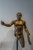 Musée d'Aquitaine. <br> Statue d’Hercule
Bordeaux, impasse Saint-Pierre en 1832 Fin du IIe siècle - tout début du IIIe siècle Alliage de cuivre
Lorsqu’elle fut découverte en 1832 dans un égout place Saint-Pierre, la statue était en plus de vingt fragments. Elle fut reconstituée une première fois en 1865 pour la XIe Exposition philomathique de Bordeaux (place des Quinconces) puis pour l’Exposition universelle de 1878 à Paris (Palais du Trocadéro). Ce n’est qu’en 1963 à la faveur d’une exposition sur l’Art dans l’Occident romain au Musée du Louvre qu’elle fut restaurée définitivement.
Malgré les parties manquantes, Hercule est identifiable à la peau de lion enroulée autour de son avant-bras gauche, celle du lion de Némée qu’il parvint à terrasser au cours du premier des « Douze Travaux » qui lui furent imposés avant d’accéder à l’immortalité. On suppose aujourd’hui qu’il soutenait sa célèbre massue de la main gauche, tandis que, de la droite, il tendait la coupe d’ambroisie, breuvage des dieux et symbole de son immortalité.
Il est représenté dans la nudité héroïque, en appui sur la jambe droite, l’autre jambe un peu fléchie en arrière. Le mouvement du corps est habilement exprimé par le déhanchement en opposition avec la ligne des épaules, et souligné par la tête légèrement tournée vers la droite. Avec cette pose très en vogue dans la sculpture grecque classique, l’influence du célèbre sculpteur grec Lysippe se lit notamment dans les proportions du corps et la musculature puissamment rendue.
Le rapprochement avec certains portraits de l’empereur Septime Sévère en Hercule conduit à voir peut-être dans la statue de bronze une représentation symbolique impériale.