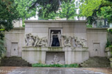 <center>Le cimetière du Père Lachaise </center> Le monument aux morts de l'ossuaire, par le peintre et sculpteur Albert Bartholomé, inauguré le 1er novembre 1899, après douze ans de travaux. À gauche, et suivant une ligne ascendante, une femme est assise, les cheveux dénoués ; derrière elle, un jeune homme, accroupi ; une femme agenouillée, de ses deux bras, se fait un bandeau pour ne pas voir la porte sombre près d'elle, la main tendrement appuyée sur son épaule, son compagnon lui donne un suprême baiser ;  appuyés l'un sur l'autre, dans un dernier groupe, le visage caché par leurs bras  ou leurs mains, deux autres malheureux attendent l'inexorable mort. Et de l'autre, côté, suivant une ligne descendante, un homme chancelant qui, de ses deux mains, s'accroche au bord de la porte dont il va franchir le seuil ; une femme, prosternée, la face contre terre, une vierge, les mains jointes, un enfant, et un peu plus loin, une femme aux longs cheveux défaits, soutenue par l'étreinte fraternelle d'un ami ; une jeune fille, au moment de s'engager sur la voie douloureuse, se détourne soudain et envoie, de ses doigts un baiser  au cher passé qu'elle quitte. Mais un couple est entré dans le caveau, et, rasant des deux côtés la paroi de l'étroit couloir, s'enfonce dans l'ombre qui déjà l'enveloppe. Au-dessous, une large baie est creusée ; un homme et une femme sont étendus côte à côte, leurs quatre mains unies et leurs visages rapprochés, un enfant est comme jeté en travers de leurs corps. Agenouillée au-dessus d'eux, une figure radieuse ouvre ses deux bras étendus.