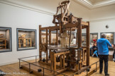 <center></center><center>Musée des Arts et Métiers.</center> Métier à tisser les façonnés de Vaucanson. Jacques Vaucanson, célèbre dans toutes les cours d’Europe pour ses automates, est nommé en juin 1741 inspecteur des manufactures de soie. il commence alors une tournée d’inspection des établissements et imagine des machines qui améliorent grâce à des dispositifs mécaniques le moulinage de la soie ou le tissage des tissus façonnés.
Cames, manivelle et vis sans fin.
Le métier combine plusieurs recherches de Vaucanson : la programmation, la reproduction du mouvement humain et un système ingénieux Bile navette. Cette dernière, entièrement métallique, est saisie alternativement par des bras munis de pinces, fixés chacun sur un chariot de bois. La mécanique, le chasse- navette et le battant sont entraînés par des cames. Le tissu s’enroule régulièrement grâce à une vis sans fin.
Un automate à tisser.
Véritable automate à tisser, le métier est entièrement mis en mouvement par une simple manivelle et transforme ainsi radicalement les gestes du tisseur. En 1747, Le Mercure de France rapporte : « On voit sur le métier l’étoffe se fabriquer sans aucun secours humain, c’est-à-dire la chaîne s’ouvrir, la navette jeter la trame, le battant frapper l’