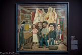<center>Les Grands Peintres Naïfs.</center> André Bauchant
1873-1968
La Boucherie
1940
Huile sur toile.
La Boucherie est une scène de vie quotidienne à Château- Renault, la ville natale de Bauchant Elle est peinte en 1940, au début des restrictions alimentaires mises en place sous l'Occupation. Le pain, le fromage et la viande sont les premières denrées concernées. Les carcasses dominent la composition. On reconnaît ensuite la bouchère près du billot, le capitaine des pompiers avec son chien, une notable discutant le prix de sa commande. Le choix du sujet et l'attention portée aux détails—les briques apparentes derrière la peinture écaillée, les maisons à colombage visibles derrière la fenêtre—respectent les codes de la peinture de genre, celle du XVIIe siècle hollandais ou encore celle des frères Le Nain que l’exposition «Les martres de la réalité» avait remis à l’honneur en 1934.
