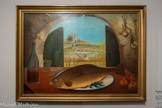 <center>Les Grands Peintres Naïfs.</center> Ferdinand Desnos
1900-1958
La Carpe
(Nature morte à la carpe)
Non daté
Huile sur bois.
Attaché à sa région natale, Ferdinand Desnos situe souvent des paysages de Touraine à l'arrière-plan de ses compositions. On reconnaît ici le pont sur le Cher et la tour carrée du château de Montrichard où le peintre a vécu avec sa famille au début des années 1920. Le caractère préservé de ce village, l'arche de pierre derrière lequel il se découpe et le choix des objets présentés au premier plan donnent à cette nature morte un caractère intemporel.
Le choix de la carpe comme objet principal du tableau est peut-être à prendre comme un symbole christique de résurrection, étant donné le caractère fréquemment religieux des œuvres de Desnos.